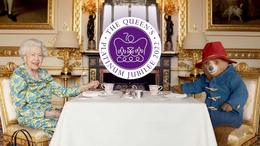 The queen and Paddington pose in an ornately gold decorated toom at a table with tea cups.