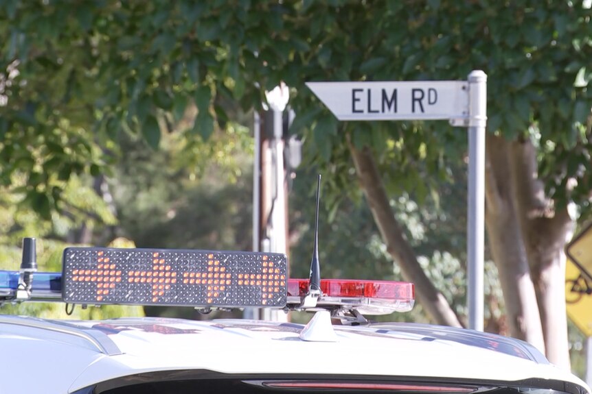 The top of a police car with arrows pointing right, in the background is a sign that reads Elm Rd