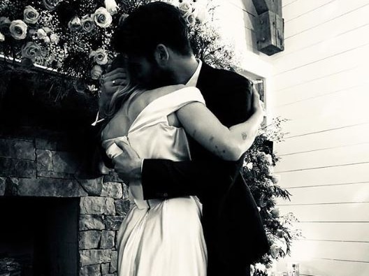 A woman in a long dress hugs a man in a suit as they stand in front of a fireplace.