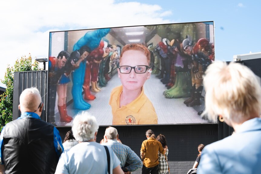 A photo of Peter Dodt is seen on a big screen at the memorial. People are looking up at it.