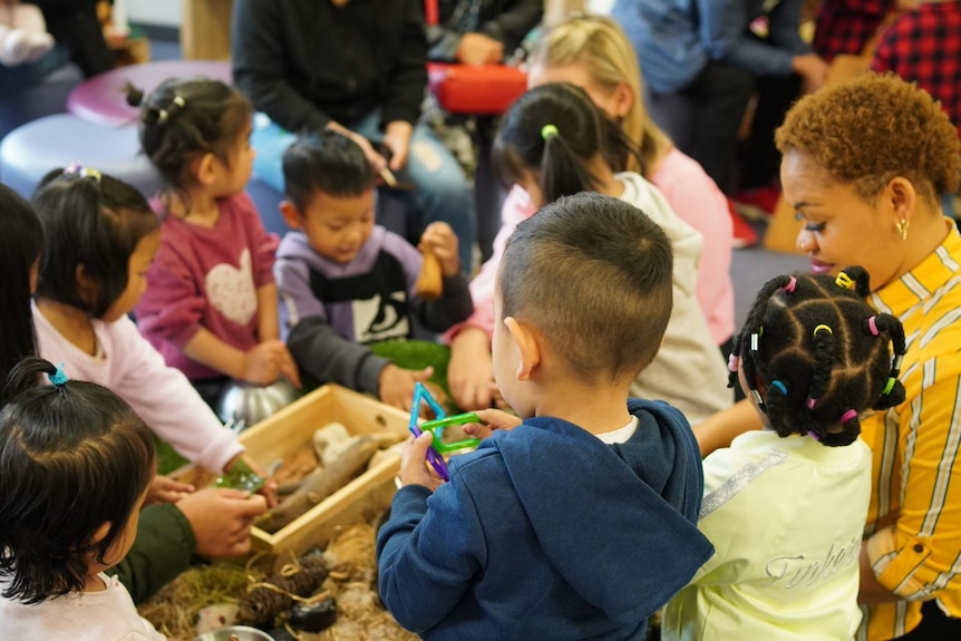 Seven young children playing with a wooden box of sand and flowers in an indoor setting