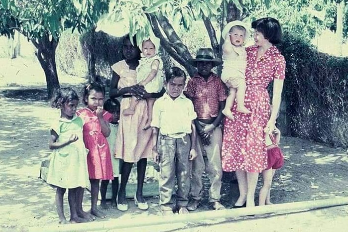Indigenous children stand under a tree on a station with an Asian woman holding a baby