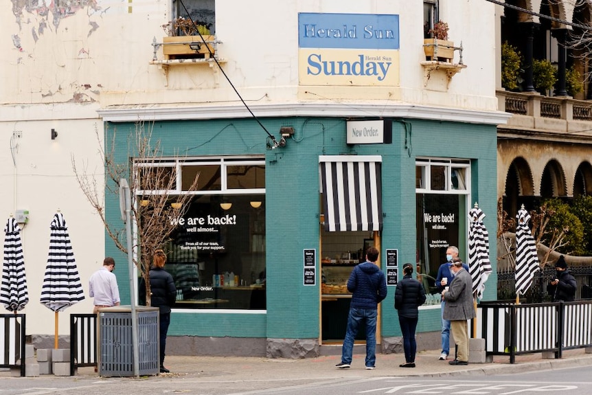 Seven people wait outside a green and white Melbourne corner cafe for takeaway during coronavirus restrictions.