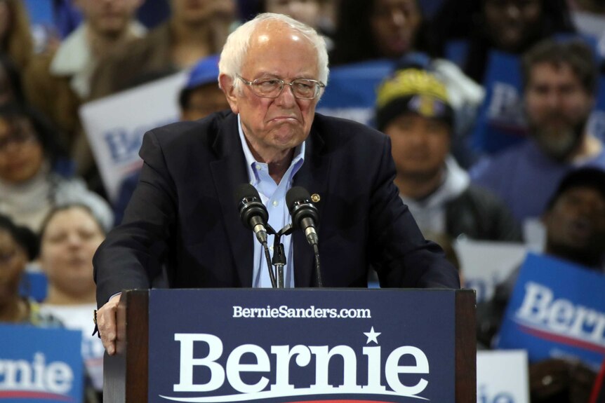 Bernie Sanders stands at a podium looking grumpy as he addresses a crowd of supporters.