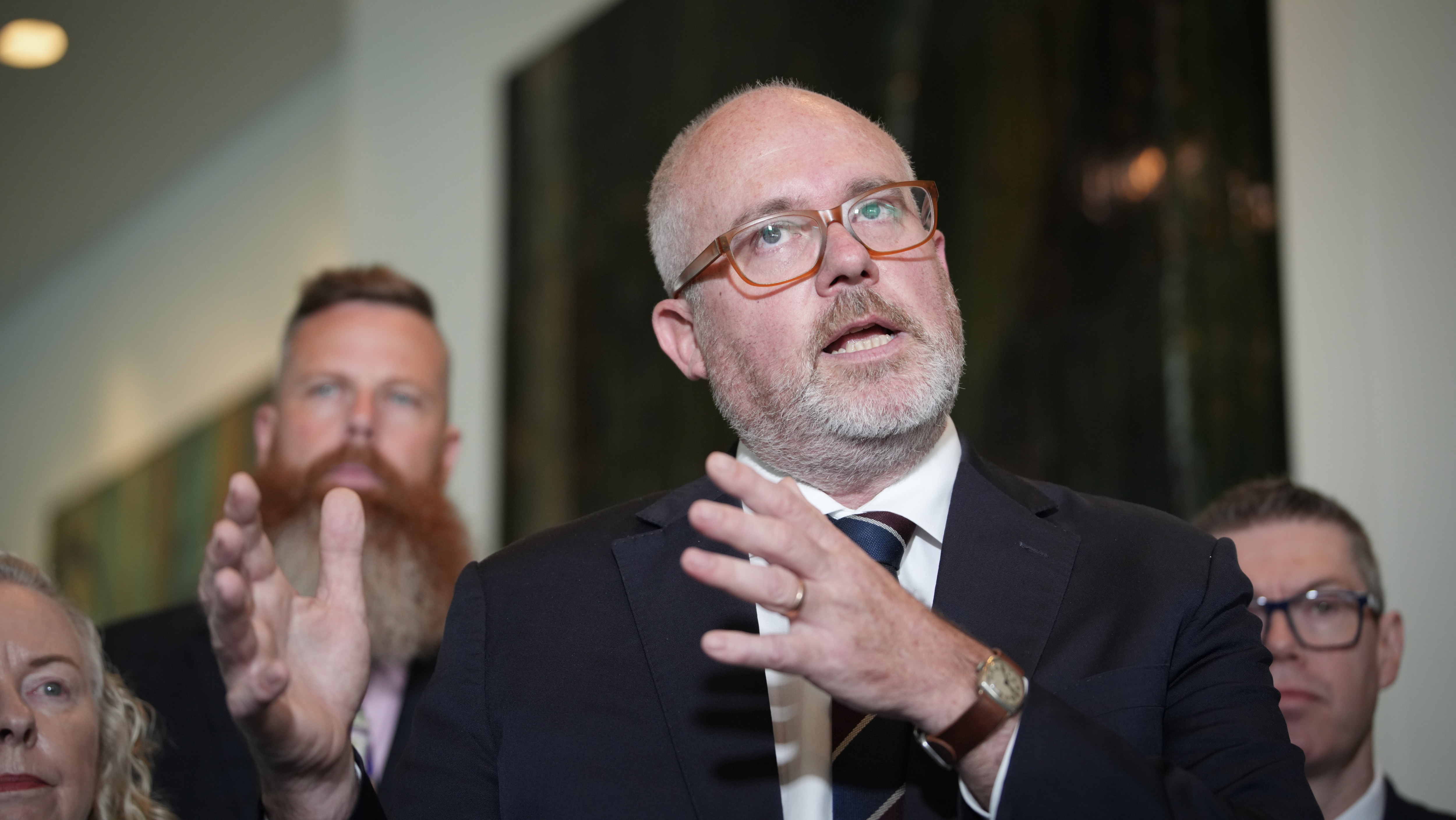 Tim Ayres está falando, tem barba por fazer branca, óculos, gravata preta e paletó. 