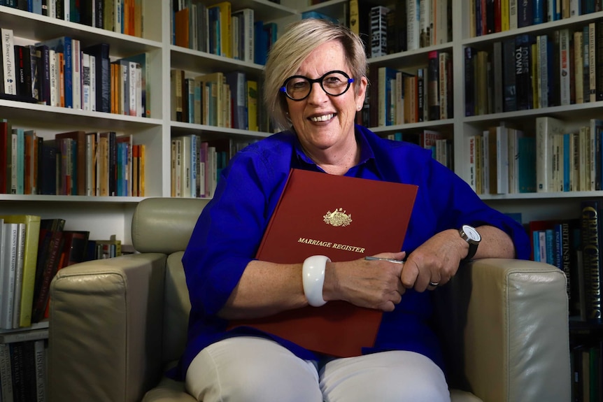 A woman sits on a chair holding a book that reads Marriage Register.