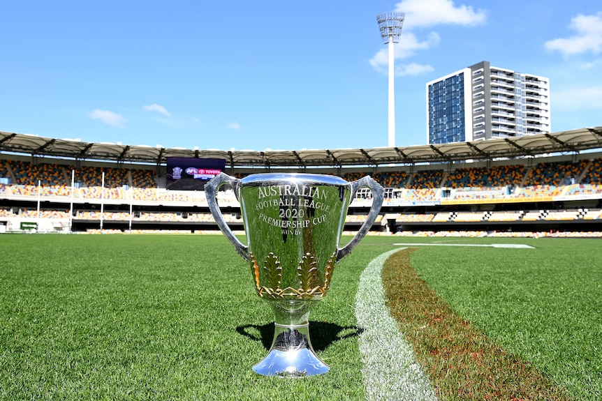 The AFL trophy sits on the grass at the Gabba with empty stands and a set of goalposts in the background.