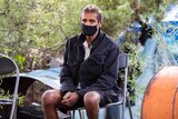 A man in shorts and a jacket, wearing a mask, sits on a chair in front of a camp hidden in bushes.