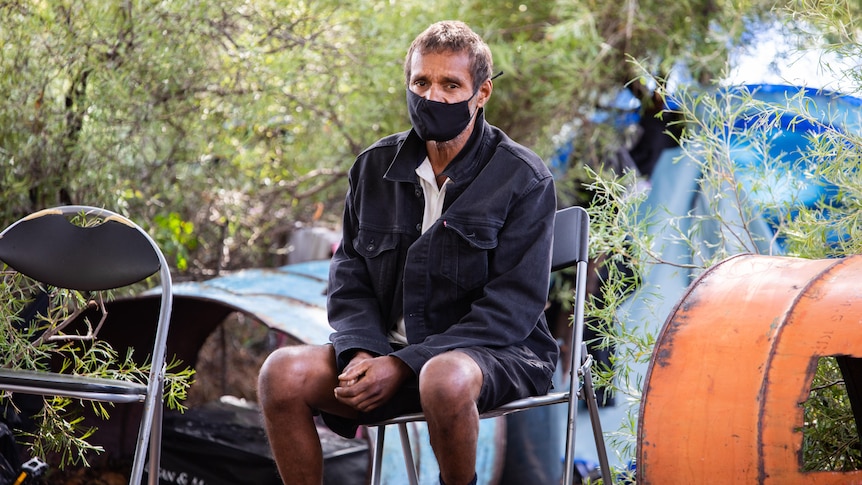 A man in shorts and a jacket, wearing a mask, sits on a chair in front of a camp hidden in bushes.