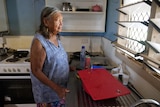 An elderly indigenous woman stands in a kitchen looking out a window. 