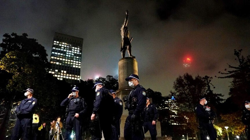 Police wearing face masks stand around a statue in a park with the skyline in the background.