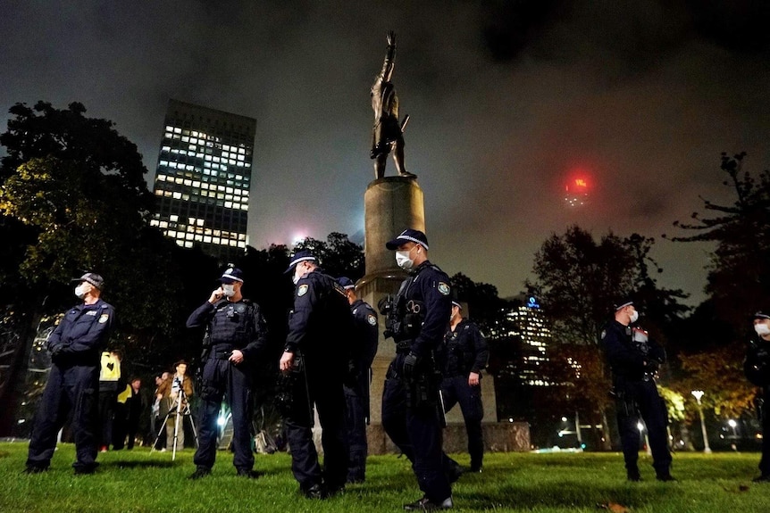 Police wearing face masks stand around a statue in a park with the skyline in the background.