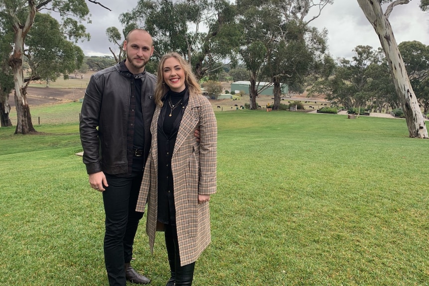 A man and woman stand next to each other in front of gum trees. 