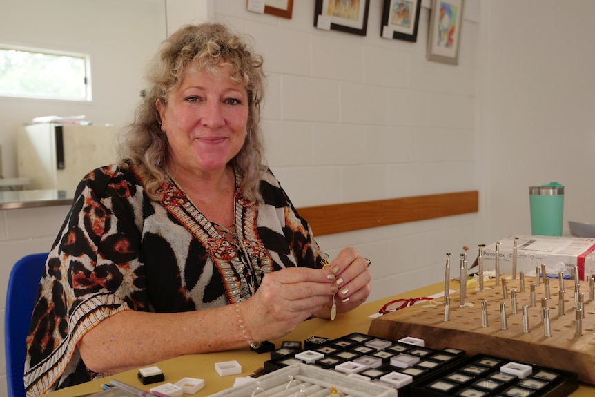 Cindy Kelly sitting behind her precious gem display, holding one, smiling at the camera, curly hair.