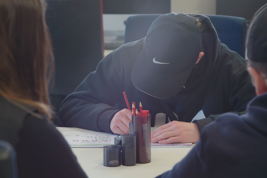A boy wearing a cap sits at a desk, writing with a pencil in a workbook.