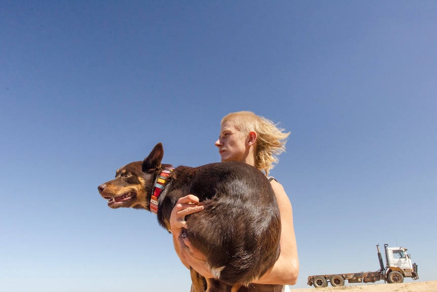 A mullet-haired shearer holding a dog against the back drop of a clear blue sky