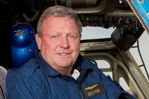 A man dressed in blue overalls sits in the cockpit of a helicopter.