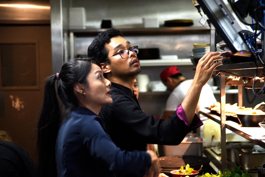 A woman and a man look seriously at a screen in a busy restaurant kitchen. 
