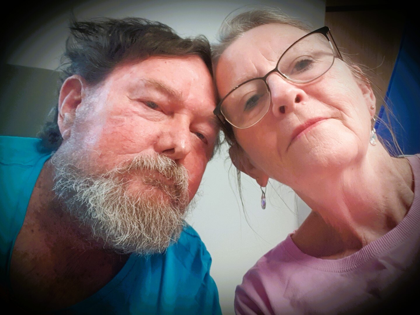 A man with a light bear and dark hair with his head nestling into a woman wearing glasses and earrings.