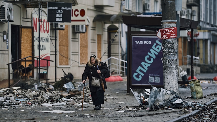 An old lady with a stick walks through a street that is covered in debris.