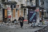 An old lady with a stick walks through a street that is covered in debris.