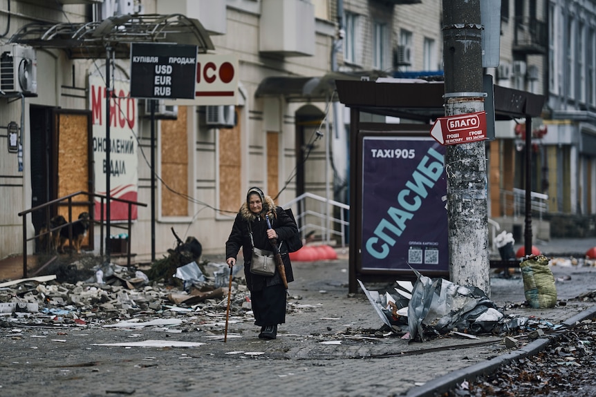 An old lady with a stick walks through a street that is covered in debris.