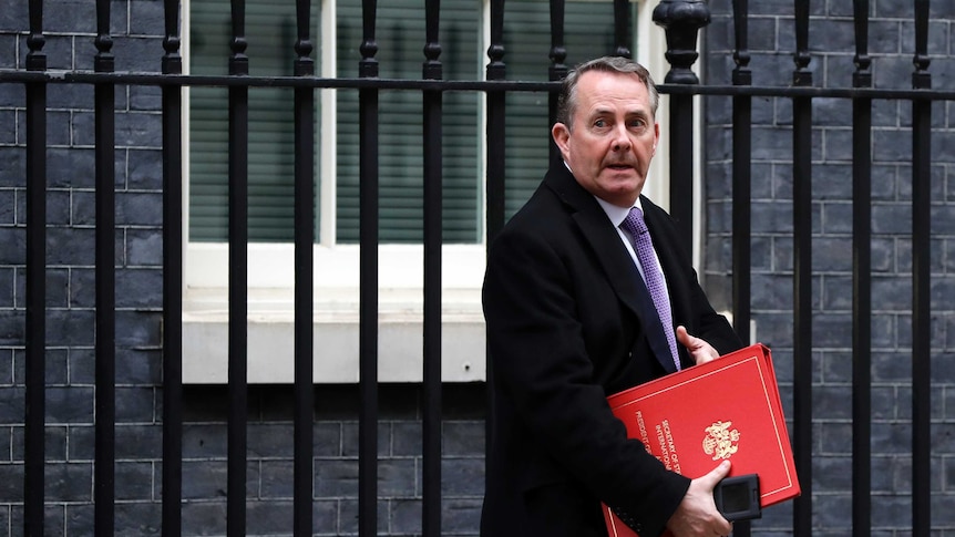 Liam Fox glances to the left of frame in front of black wrought-iron gates in front of a Georgian bluestone house.