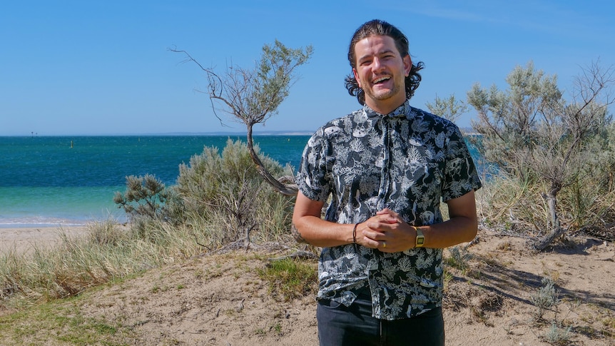 Bransen Gibson standing on a beach dune, with the ocean and bright blue sky behind him.