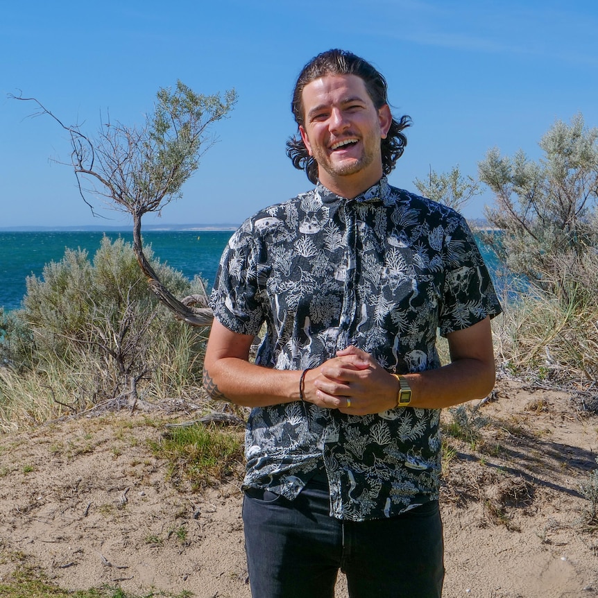 Bransen Gibson standing on a beach dune, with the ocean and bright blue sky behind him.