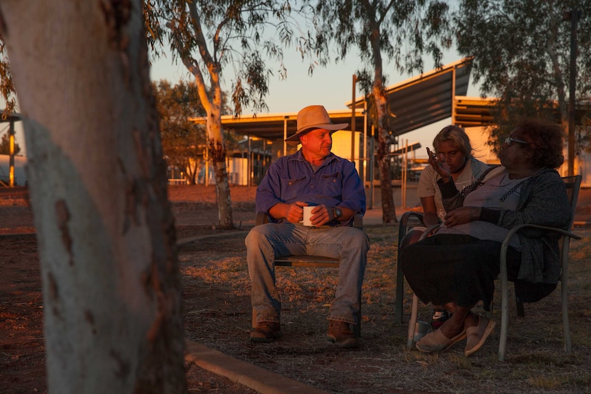 A man and two women sit outside, under trees, with a cup of tea