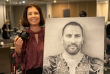 A woman in a red shirt holding a camera, standing near a black and white portrait of a tattooed man
