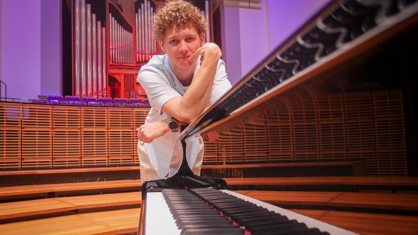 A young man with curly hair leans against a piano.