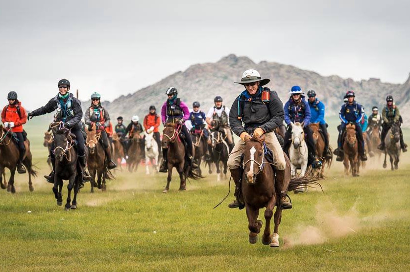 A large group of horse riders gallop across a grassy plain with hills in the background