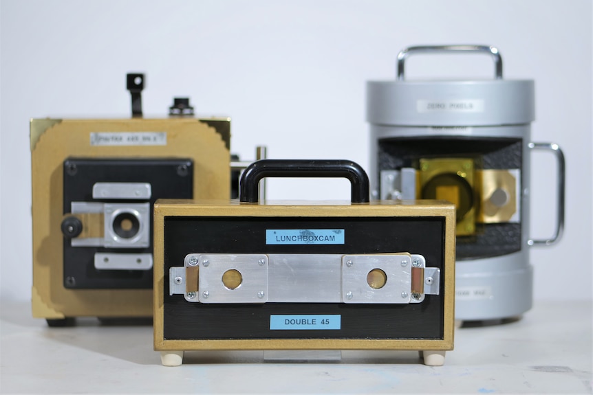 Three different pinhole cameras, with metal 'lunchbox camera' in front and cardboard version behind at left