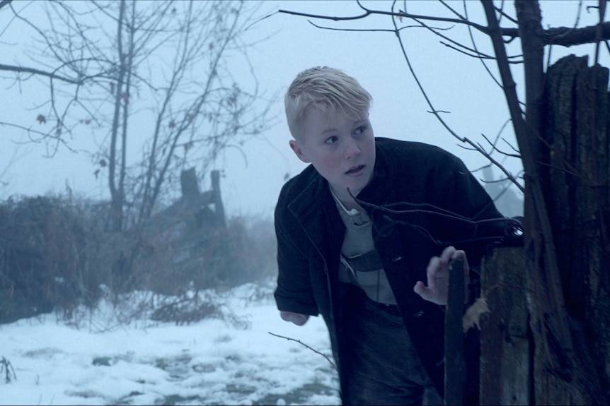 Knox wearing a black coat, peering around a wooden structure in a snowy landscape.