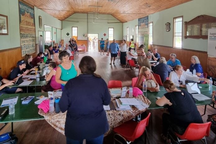 a wide shot of a community meeting in a hall, people at tables and standing around