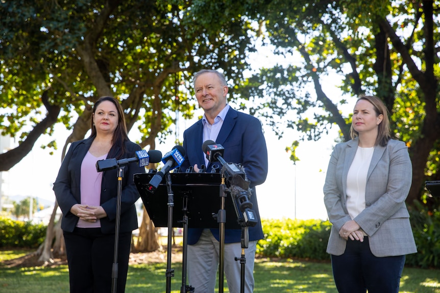 A man speaks at a group of microphones. He is flanked by two women, one to his left and the other his right.