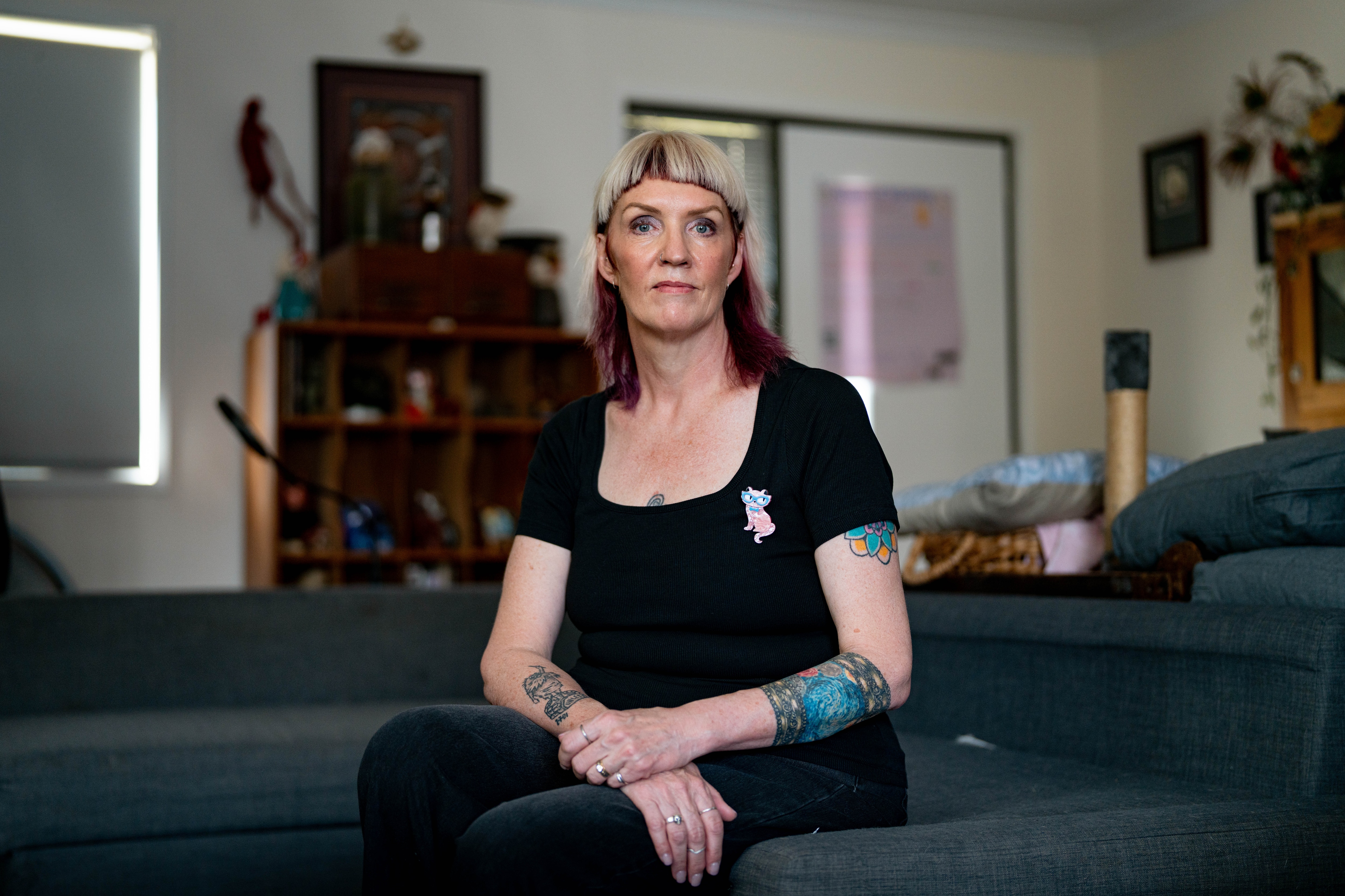 Wide shot of Leonie, wearing a black t-shirt with a pink cat broch, sitting on a couch, turned slightly, serious face.