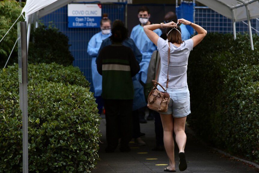 A woman walks towards the entrace of the Royal Perth Hospital COVID-19 clinic with other people in line and medics wearing PPE.