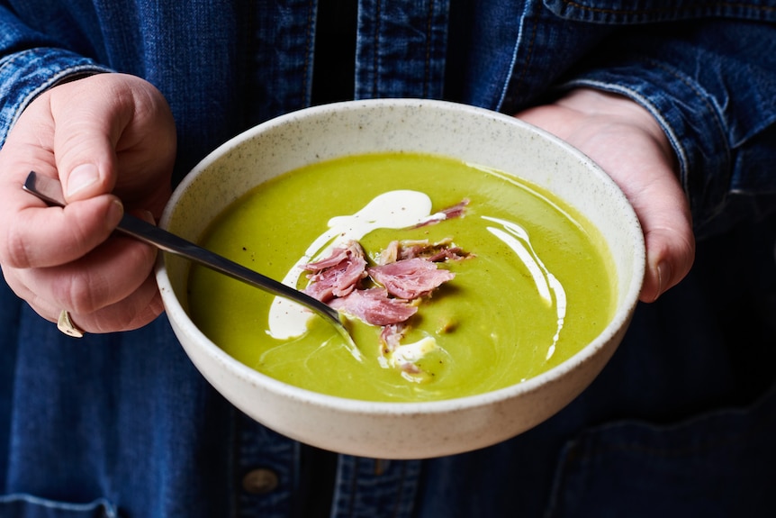 A person holds a bowl of bright green soup with pieces of ham on top and a drizzle of cream.