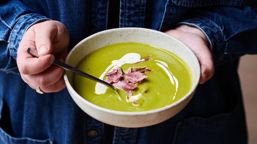 A person holds a bowl of bright green soup with pieces of ham on top and a drizzle of cream.