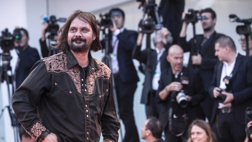 Thornton wears a black embroidered shirt and stands in front of a block of suited paparazzi.