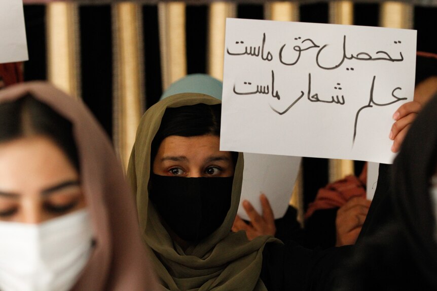 머리 스카프와 마스크를 쓴 세 명의 여성이 항의 표지판을 들고 있습니다.