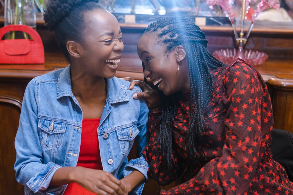 Taku and her friend sit facing each other and laughing. Taku's hand is on her friend's shoulder.