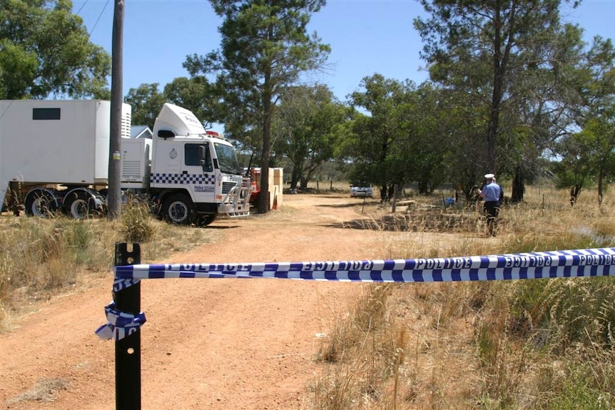 Police tape is wrapped around a steel post, with a police truck and officers in the background among trees.