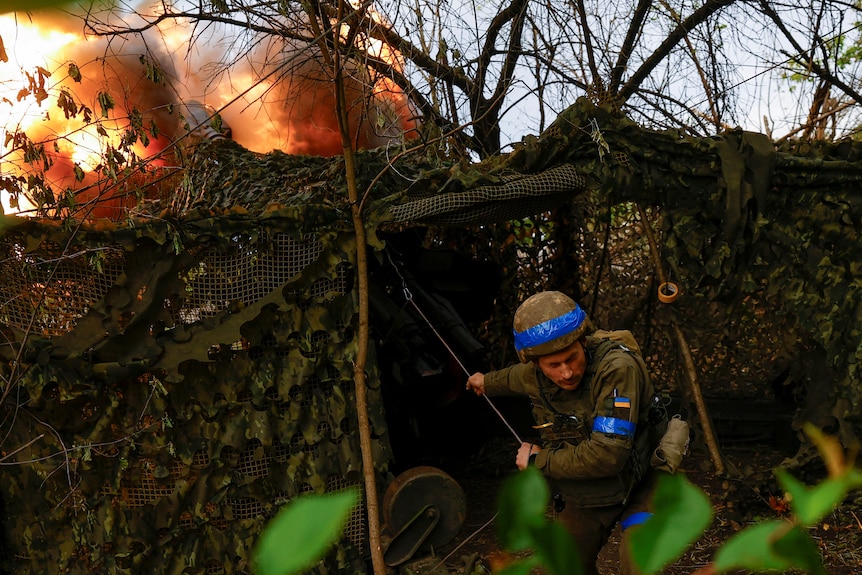 A soldier hidden behind netting crouches beneath a huge explosion 