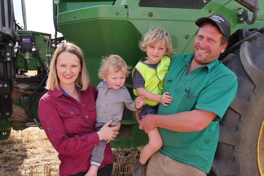 a woman and her husband hold small children, standing in front of a green tractor