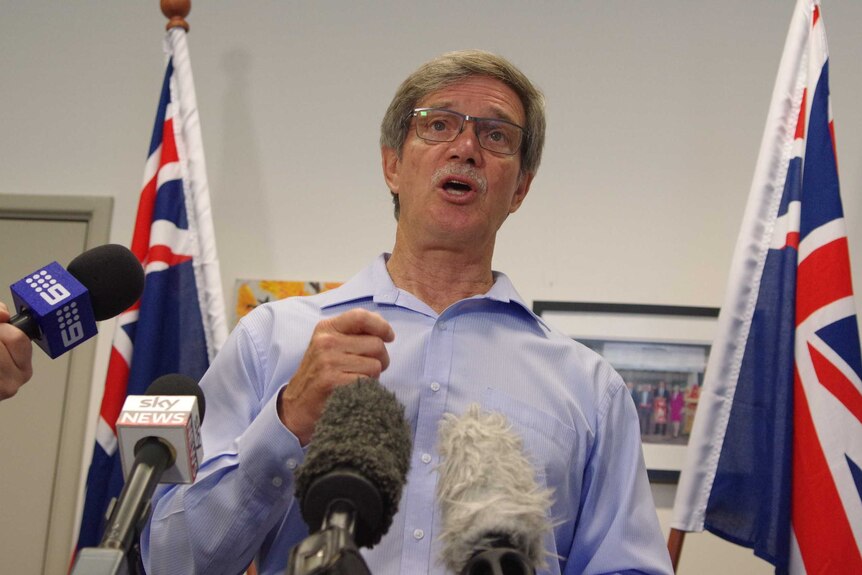 Mike Nahan in front of microphones, flanked by flags.