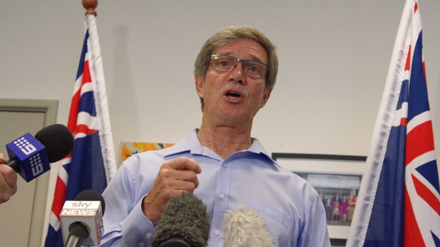 Mike Nahan in front of microphones, flanked by flags.