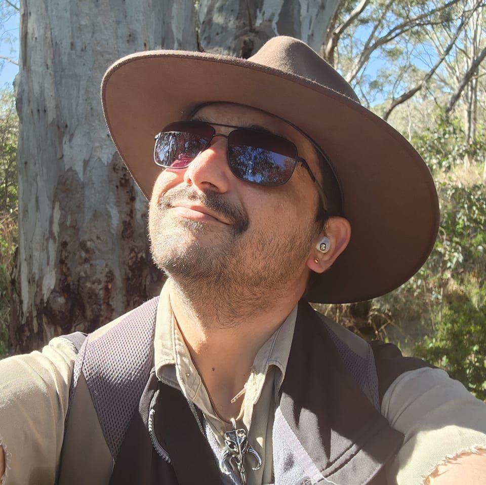 An Indigenous man wearing sunglasses and a hat, looking up at a tree in scrubland.